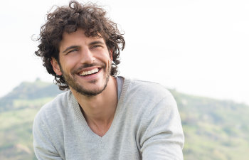 A man with curly hair and a beard smiles outdoors, wearing a light gray sweater with a blurred green landscape in the background.