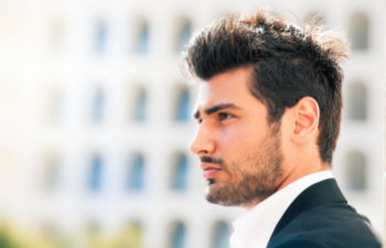 A man with dark hair and a beard, wearing a black suit jacket and white shirt, is looking to the side with a blurred building in the background.