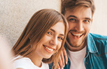 Two young adults smiling and posing for a selfie against a light-colored wall.