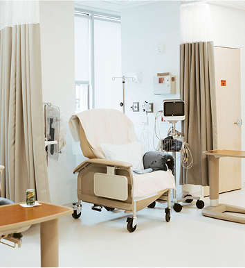 Hospital room with a recliner chair, IV stand, medical equipment, and privacy curtains. A can of soda is on a tray table beside the chair.