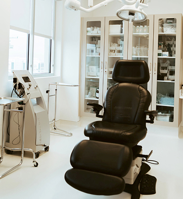 A medical examination room with a black adjustable chair, cabinets with supplies, and medical equipment in a bright, white setting.