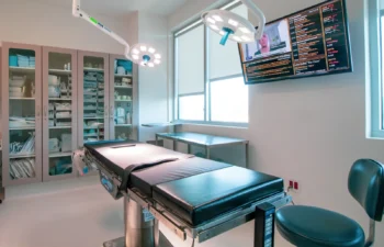 A modern, well-lit medical examination room with a padded exam table, surgical lights, medical supplies cabinet, and a large wall-mounted monitor displaying information.
