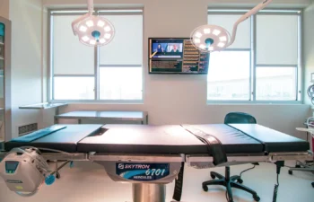 A modern surgical room with an empty operating table, overhead surgical lights, medical equipment, and a wall-mounted monitor displaying information.