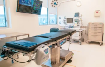 A modern operating room with an empty surgical table, medical equipment, monitors, and a wall-mounted TV displaying news.