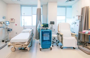 A hospital room with a hospital bed, a medical cart, monitoring equipment, and a reclining chair next to large windows with natural light.