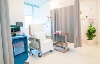 A hospital room with a medical recliner chair, side table, equipment cart, privacy curtain, and a potted plant near a window.