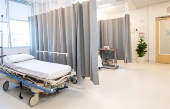 A hospital room with a wheeled bed, privacy curtains, a side table, a plant, and a closed door with an exit sign.