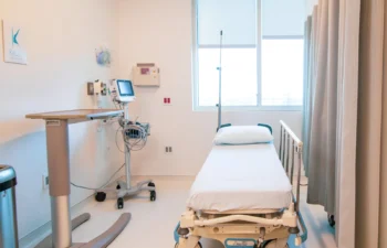 A hospital room with a single empty bed, a bedside table, medical equipment, and large windows letting in natural light.