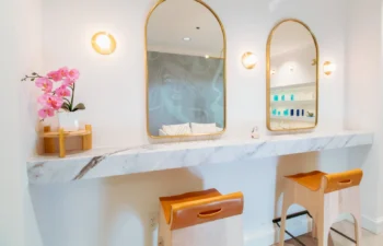 A modern vanity area with two mirrors, marble countertop, wooden stools, wall lights, and a potted pink orchid on the left.