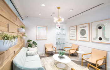 Bright, modern waiting room with tan chairs, a light gray sofa, wall art, potted plants, shelving with glass decor, and a round white coffee table under a modern light fixture.
