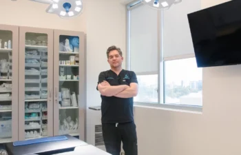 A person in medical scrubs stands with arms crossed in a well-lit clinic room, next to a treatment bed, cabinets with supplies, and medical lights overhead.
