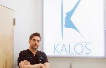 A man in black medical scrubs stands with arms crossed in front of a white wall displaying a lit-up sign with the blue Kalos logo and text.