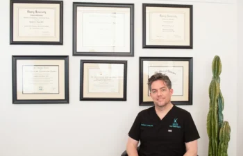 A man in black medical scrubs sits in an office with six framed diplomas on the wall and a tall cactus beside him.