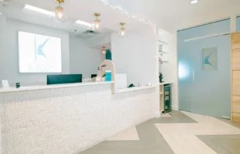 Reception area of a modern clinic with a white textured front desk, overhead lights, a logo on the wall, and glass door with a frosted logo.
