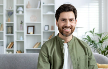 Man with dark hair and beard, wearing a green shirt, sits on a couch and smiles. Bookshelves and plants are visible in the background of a well-lit room.