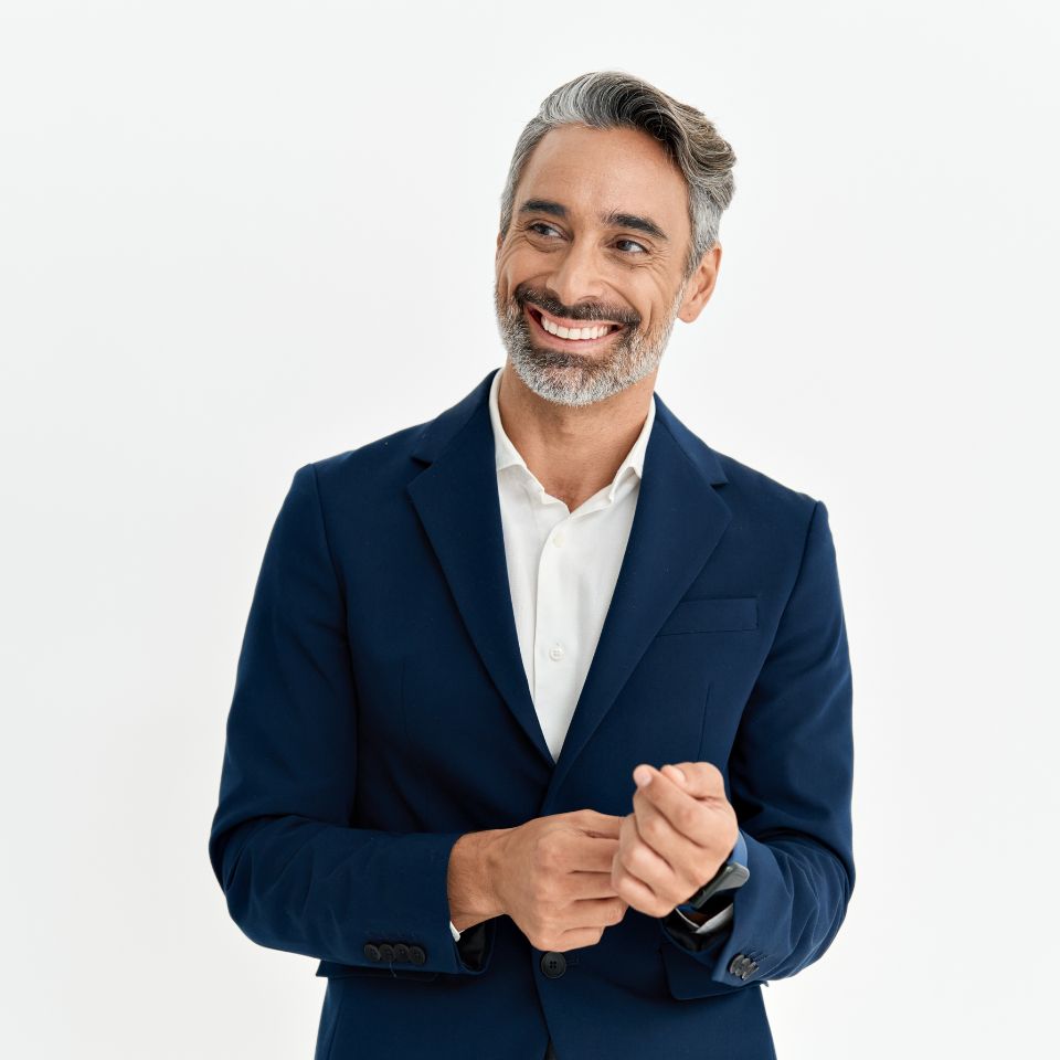A man with salt-and-pepper hair and beard, wearing a navy suit and white shirt, stands smiling against a plain white background.