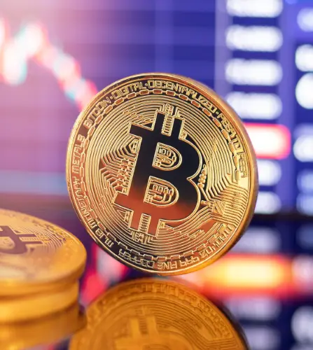 A gold-colored physical Bitcoin coin stands upright in front of a blurred background showing financial charts and data.