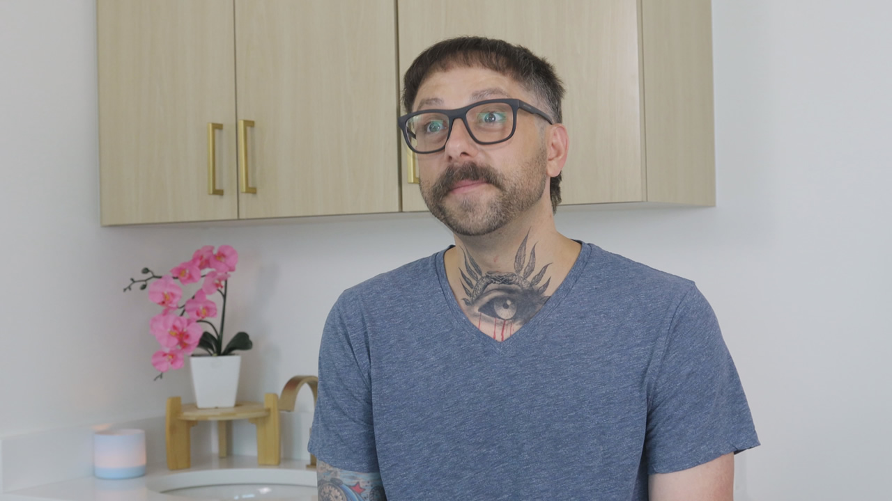 A person with glasses, a tattoo of an eye on their neck, and a mustache sits in a light-colored room with cabinets and a pink orchid plant in the background.