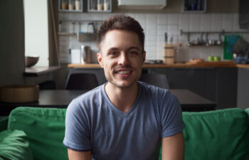 A man with short brown hair and a trimmed beard sits on a green couch, smiling at the camera in a modern kitchen setting.