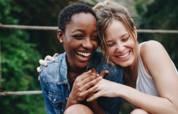 Two women sit close together outdoors, smiling and laughing with their arms around each other, with greenery in the background.