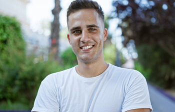 A man in a white t-shirt smiles outdoors, with greenery and trees in the background.