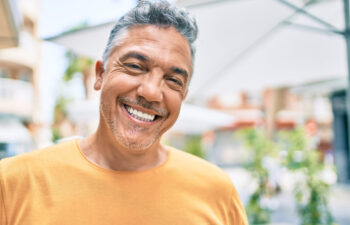 A man with gray hair and a beard, wearing a yellow shirt, smiles outdoors with blurred greenery and umbrellas in the background.