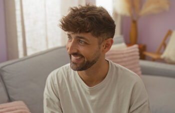 A man with short brown hair and a beard is sitting on a sofa, smiling and looking to the left in a softly lit living room.