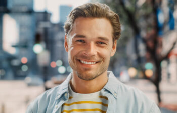 A young man with light brown hair and a beard smiles at the camera, standing outdoors on a city street with blurred buildings and trees in the background.