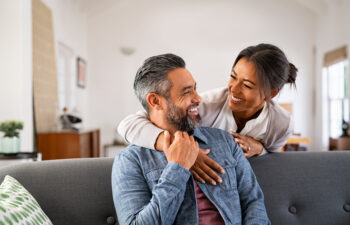 Woman standing behind a seated man, both smiling at each other in a bright living room.