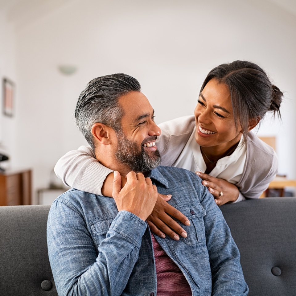 Smiling ethnic woman hugging her husband on the couch from behind in the living room. Middle eastern man having fun with his beautiful young wife on the couch. Mid adult indian man with latin woman.
