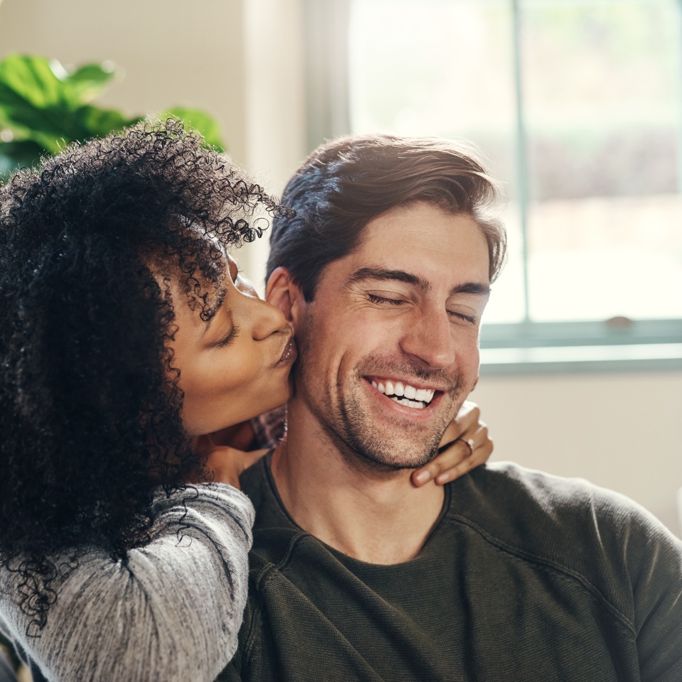 Happy man, woman and kiss on cheek on sofa for love, affection and proud for engagement. Interracial couple, hug and commitment in relationship with good news, trust and support at home with care