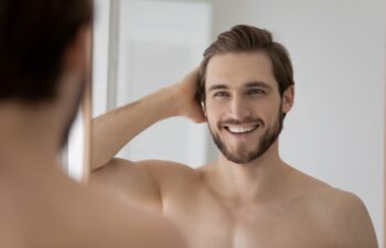 Handsome shirtless man admires his reflection in mirror, feels satisfied with quality of skin, shiny hair after morning routine of personal grooming. Cosmetics for men, skincare and haircare concept