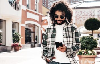Handsome smiling hipster model. Sexy unshaven Arabian man dressed in summer clothes. Fashion male with long curly hairstyle using smartphone apps, looking at cellphone screen. Holds phone